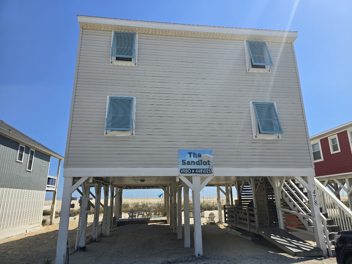 Elevated beach house with beige siding and blue shutters under a clear blue sky.
