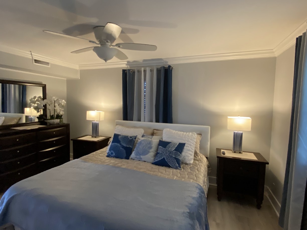 Features a bed with blue and white pillows, wood nightstands with lamps, and a dresser with flowers.