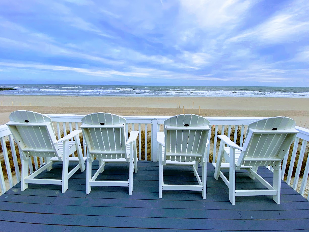 Four white Adirondack chairs face the ocean on a wooden deck,